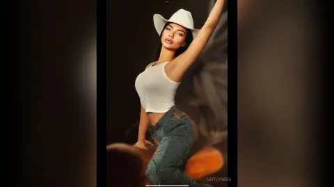 Stylish woman in white tank top and denim jeans wearing a white cowboy hat in a moody studio setting.