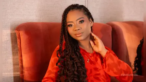 Smiling woman with long dark braids wearing a red blouse sitting on a terracotta velvet sofa in warm lighting.