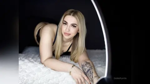 ZaraPlaya poses on a white textured bed, wearing a black bra and showing off her long blonde hair and arm tattoo.