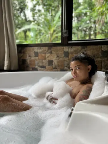 Relaxed woman relaxing in a white bubble bath in a bright room with a view of lush green trees.