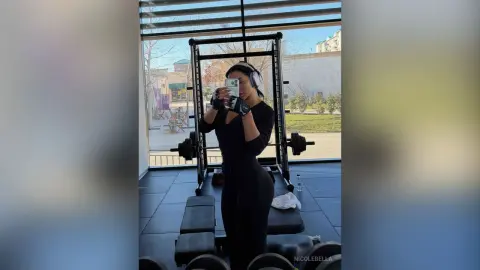 NicoleBella takes a mirror selfie in a black long-sleeve bodysuit while wearing white headphones in a gym setting.