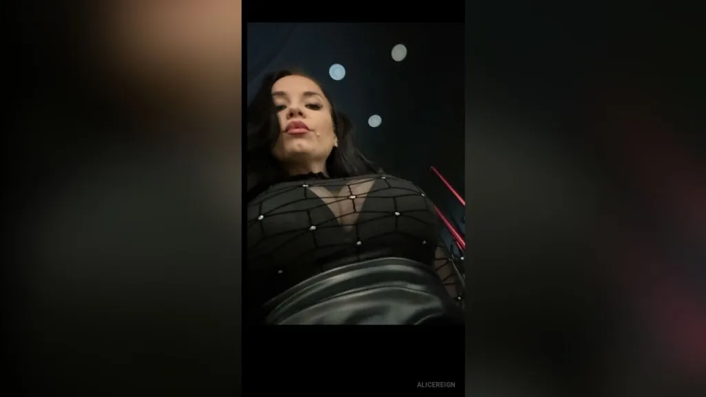 AliceReign — AliceReign wears a black sheer mesh top while looking down at the camera in a dark room.