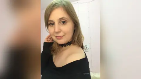 Smiling blonde woman in a black off-the-shoulder top and lace choker posing in a bright indoor setting.