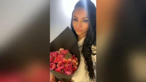 Smiling woman in a white top holding a large bouquet of pink roses in a bright indoor setting.