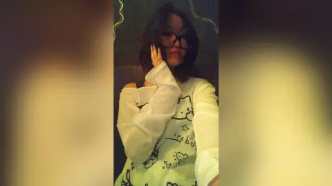 TishaOyervides wears glasses and a white long-sleeved graphic shirt while posing with one hand up to her face.