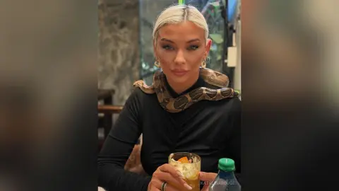 Blonde woman in a black turtleneck and snake print scarf holding a cocktail in a moody restaurant setting.