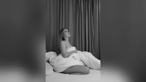 Dreamy black and white portrait of a woman relaxing under white bedding in a cozy bedroom setting.