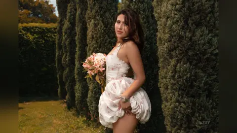 Woman in a white floral sundress holding a bouquet in a lush green garden during golden hour.