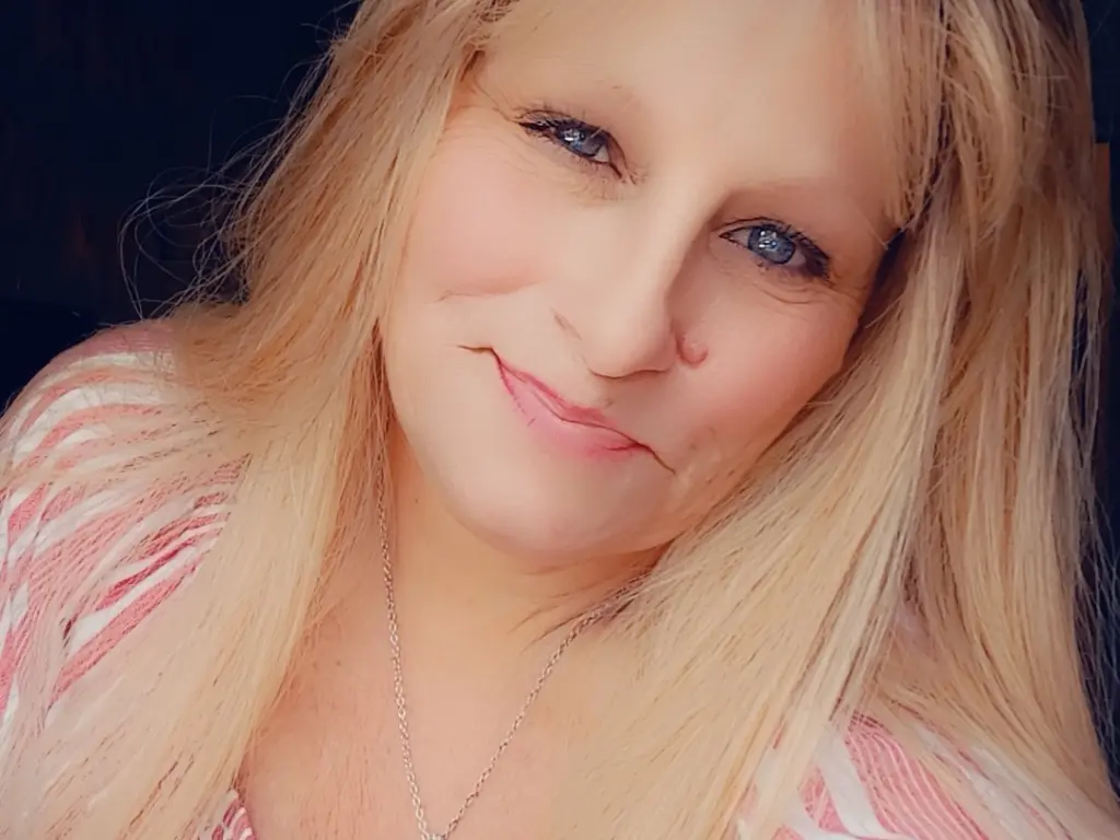 SxySusan poses with long blonde hair, wearing a white tank top that shows her cleavage and bare shoulders.