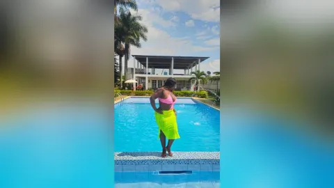 KattyLeya — KattyLeya stands by a swimming pool wearing a pink bikini top and a bright neon yellow sarong.