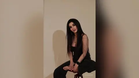 JuliDeming sits on a stool in a black tank top and black pants, posing with her long dark hair against a plain wall.