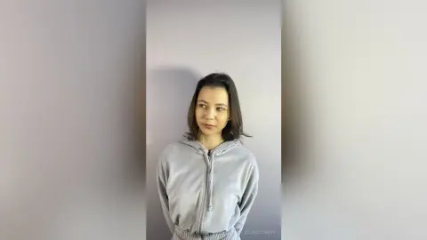 Young woman in a cozy grey zip-up hoodie posing against a minimalist neutral background with a calm expression.