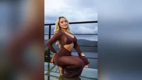 Smiling blonde woman in a brown cutout maxi dress posing on a balcony overlooking a scenic lake at dusk.