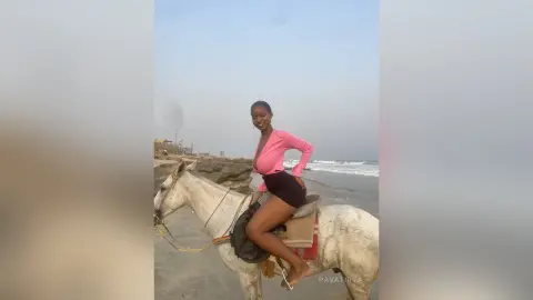 Smiling woman in a pink top and black shorts riding a white horse on a cloudy beach at sunset.