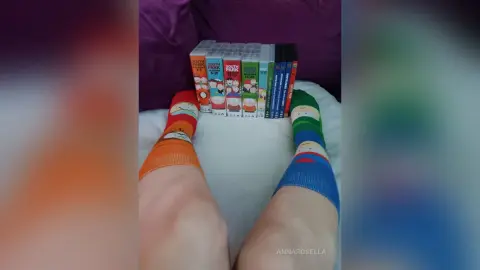 Person wearing colorful South Park character socks relaxing on a bed next to a complete DVD series collection.