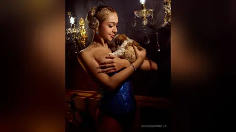 SashaNavikova wears a blue lace bodysuit while holding a small puppy against her chest in front of a lit chandelier.