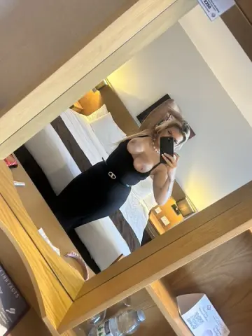 BarbieGirlGoneBad takes a mirror selfie in a black dress, showing her bare breasts and blonde hair while posing in a hotel room.