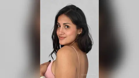 JulianaNavarro poses sideways, looking over her shoulder with long dark hair and a pink bra or top against a plain white background.
