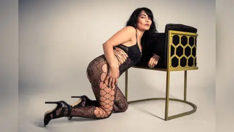 Elegant woman in black lace bodysuit and high heels posing on a gold chair in a minimalist studio setting.