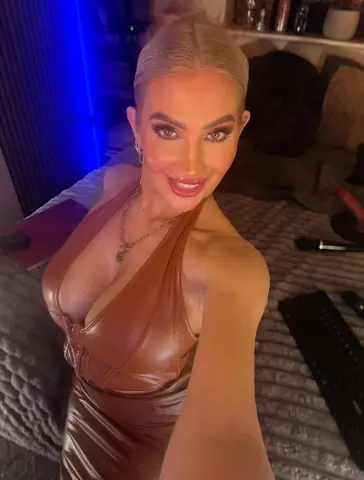 Lettie_Looner poses in a brown leather-style dress, showing off her cleavage and blonde hair in a dimly lit room.