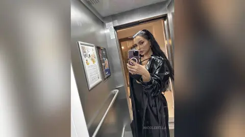 NicoleBella poses in a mirror selfie wearing a black shiny PVC or leather outfit inside an elevator.
