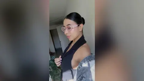 KimberlyIsaza wears pink glasses and a black sleeveless top, posing with her dark hair pulled back in a bun against a plain wall.