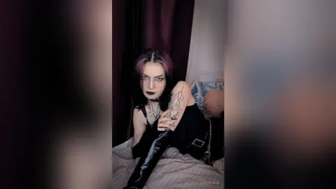 LorenLoiss poses on a bed wearing black lace lingerie and a choker, looking at the camera with dark lipstick and purple-tinted hair.