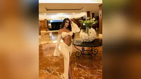 CristalMatthews poses in a white off-the-shoulder crop top and matching skirt standing next to a glass table in a marble hallway.