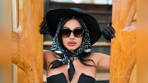 Stylish woman in a black hat and patterned headscarf posing between wooden pillars in a chic outdoor setting.