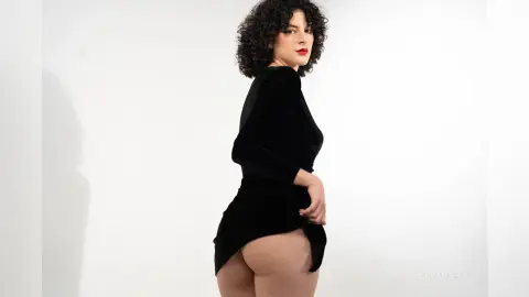RoxyGraff poses in a black long-sleeved dress, looking back over her shoulder while showing her bare buttocks against a white background.