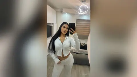 Young woman with long dark hair wearing a grey lounge set taking a mirror selfie in a modern bright room.