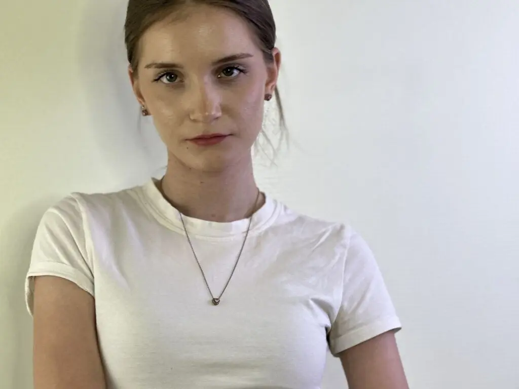 SibylMuratalla looks directly at the camera wearing a white t-shirt and a small heart pendant necklace against a plain white wall.