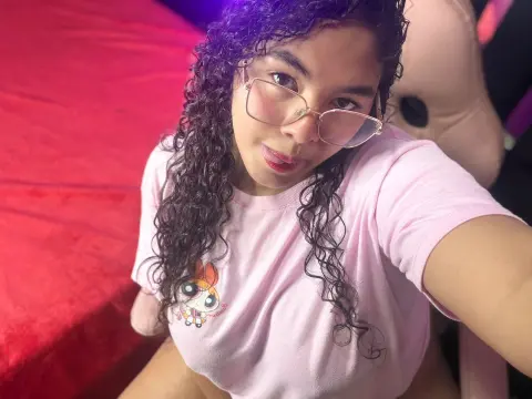 AshleyJeneer wearing glasses and a pink t-shirt with a cartoon graphic, posing with her long curly hair down.