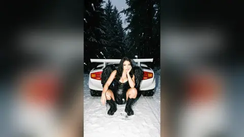 Gothic woman in black outfit and angel wings squatting in front of a white car in a snowy forest at night.