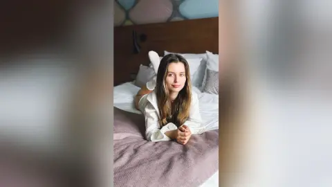 Young woman in white loungewear relaxing on a bed in a bright, cozy bedroom setting.