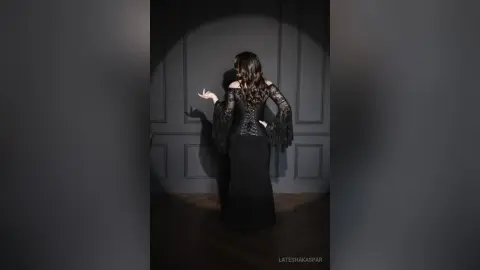 LateshaKaspar poses with her back turned, wearing a long black lace dress with flared sleeves against a dark paneled wall.