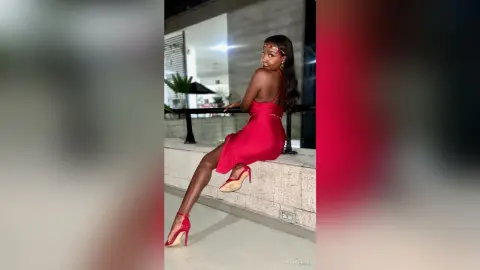 Elegant woman in a red cocktail dress and high heels sitting on a balcony at night.