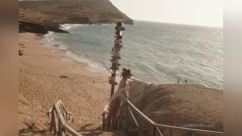 LaurentBelluci stands on a wooden balcony overlooking a sandy beach and ocean, wearing a white patterned bikini.