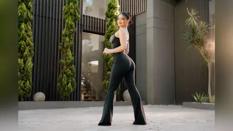 Stylish woman in black jumpsuit posing outdoors near modern architecture and greenery.
