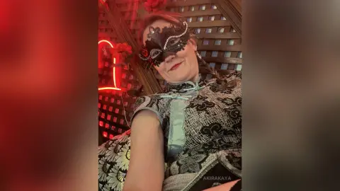 Woman in a black lace masquerade mask and patterned silver dress posing in moody red lighting.