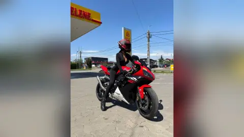 Woman in black riding gear on a red and white sport motorcycle at a gas station on a sunny day.