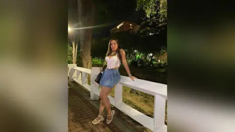 Young woman in a white lace top and denim skirt posing by a white fence at night.
