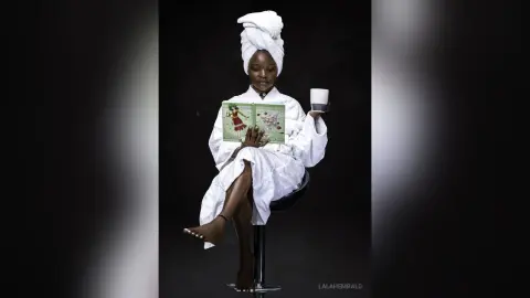 Young Black woman in a white bathrobe and towel turban reading a book with a cup in a cozy, relaxing mood.