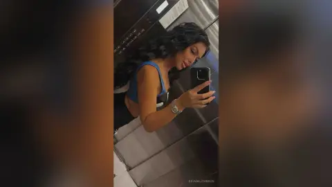 Young woman in a blue crop top taking a mirror selfie in a modern elevator.