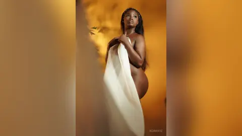 KeianHoz poses sideways against a warm gold background, partially covering her bare breasts with a white cloth.