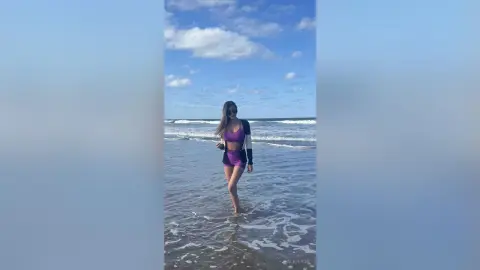 Stylish woman in a purple bikini and black cardigan walking on a sunny beach under a blue sky.