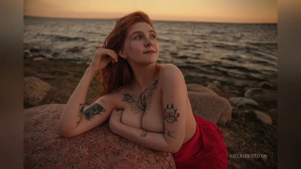 MelanieStoyn โ Redheaded woman with tattoos relaxing on coastal rocks during a peaceful sunset at the beach.