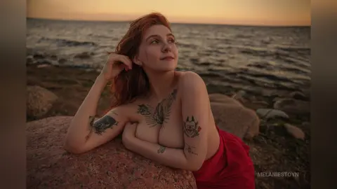 Redheaded woman with tattoos relaxing on coastal rocks during a peaceful sunset at the beach.