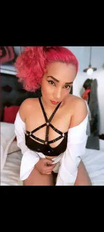 Woman with pink hair wearing a black strappy bra and white shirt sitting on a bed in a moody indoor setting.