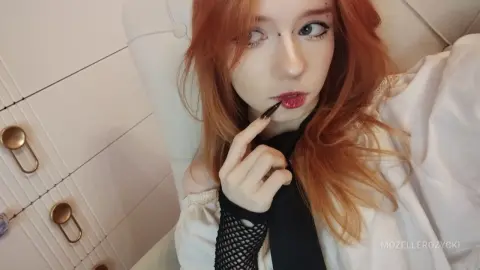 Redheaded woman in a white blouse and black tie posing with a moody expression on a white sofa.